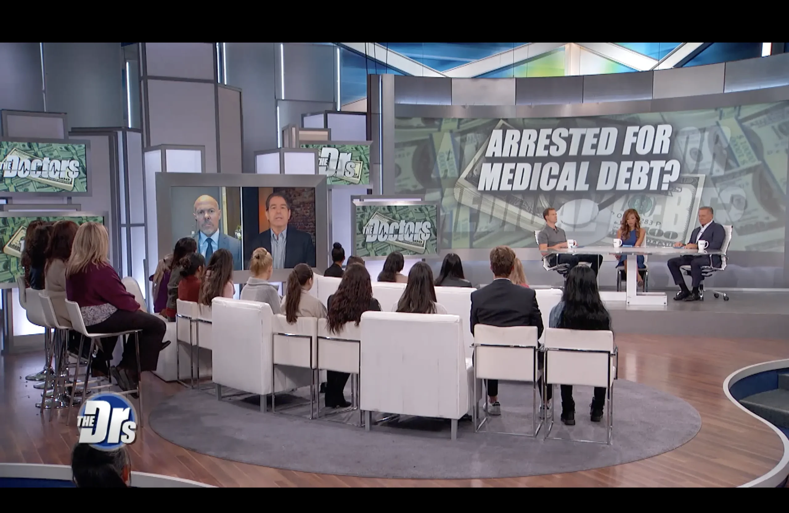 Television studio set of "The Doctors" with audience, hosts, and large screen reading "Arrested for Medical Debt?