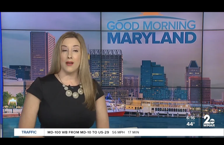 Television news anchor delivering "Good Morning Maryland" with city skyline and traffic update.