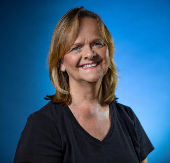 Portrait of a middle-aged woman with blonde hair wearing a black shirt against a blue background.