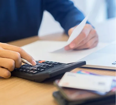 Person using a black calculator and holding a paper document on a wooden desk.