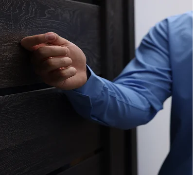 Person wearing a blue shirt knocking on a dark wooden door with a closed fist.