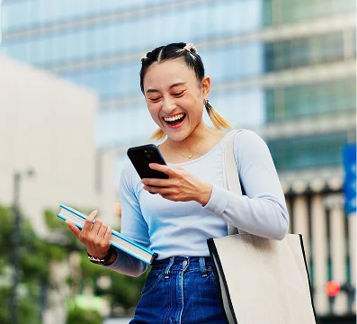 Young woman in light blue top and jeans smiling while looking at smartphone outdoors.
