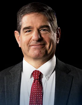 Portrait of a middle-aged man in a dark suit, white shirt, and red patterned tie against a black background.