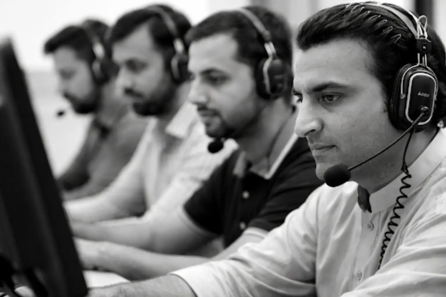 Black and white image of four men wearing headsets, working at computer stations in an office.