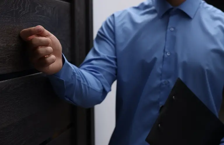 Man in blue shirt knocking on dark wooden door, holding black folder.