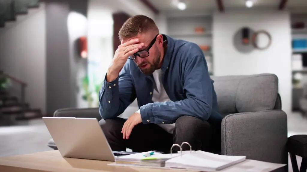 Man wearing glasses and blue shirt appears stressed, sitting on gray sofa with laptop and documents on table.