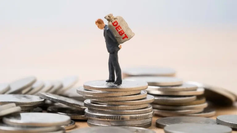Miniature figure of a man carrying a burlap sack labeled "DEBT" standing on stacked silver coins.