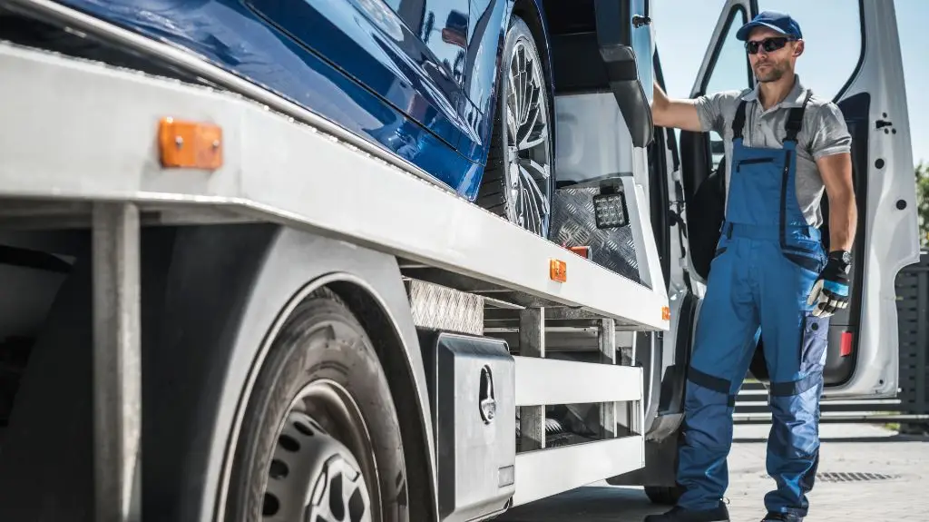 Man in blue overalls and cap standing next to a blue car on a white flatbed tow truck.