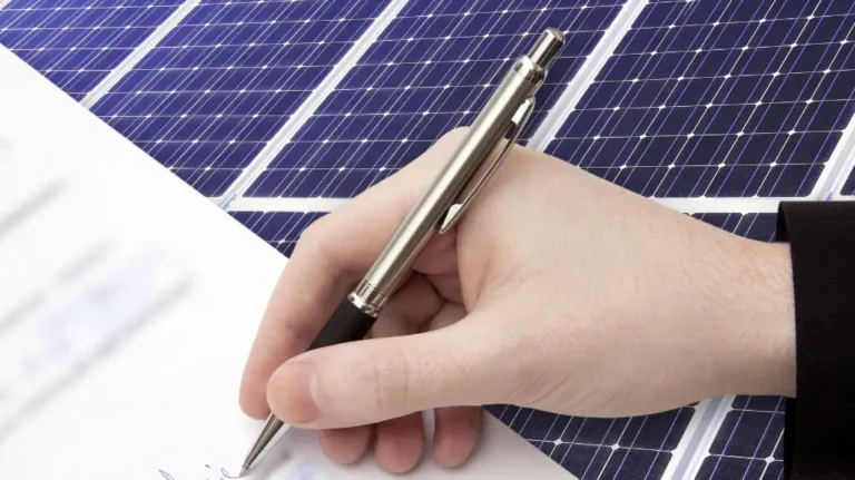 Close-up of a hand signing a document with solar panels in the background.