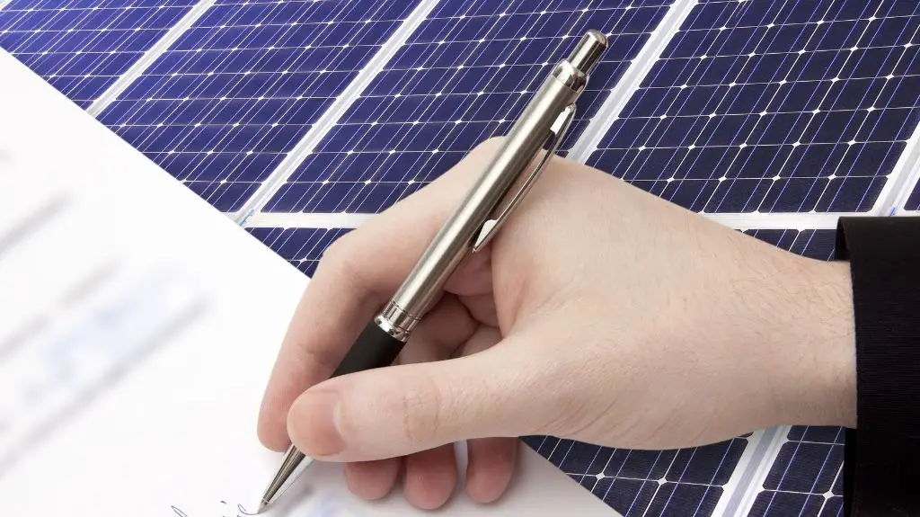 Close-up of a hand signing a document with solar panels in the background.