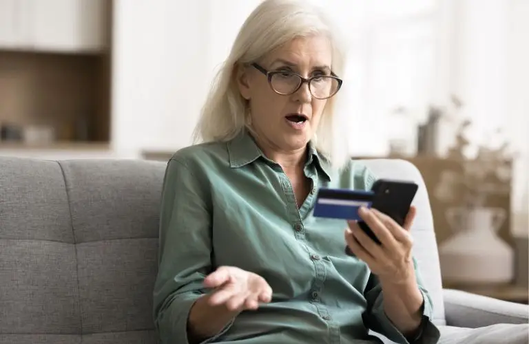 Middle-aged woman with glasses sitting on gray couch, holding credit card and smartphone, looking surprised.