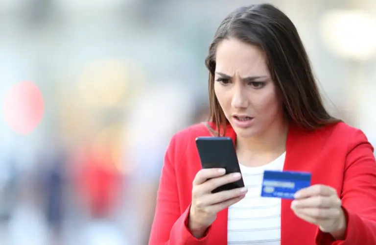 Woman in red blazer looking concerned at smartphone while holding a blue credit card.