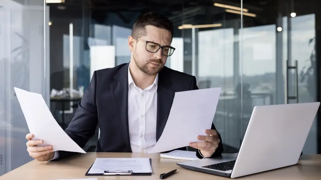 Professional man in dark suit and glasses reviewing documents at office desk with laptop.
