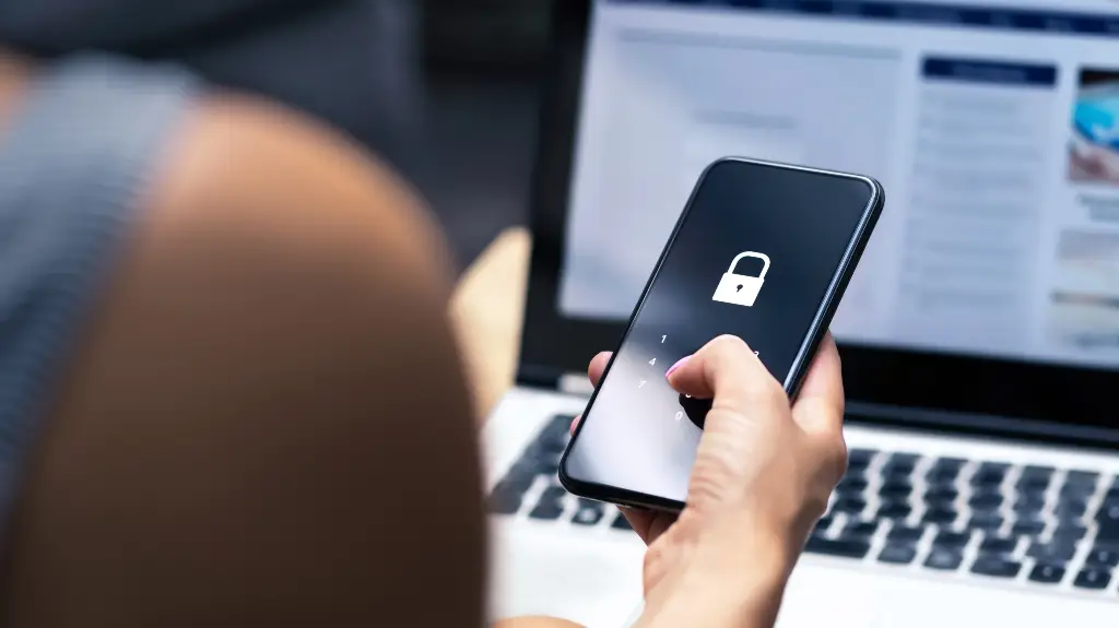 Person holding smartphone displaying locked screen with passcode interface in front of laptop keyboard.