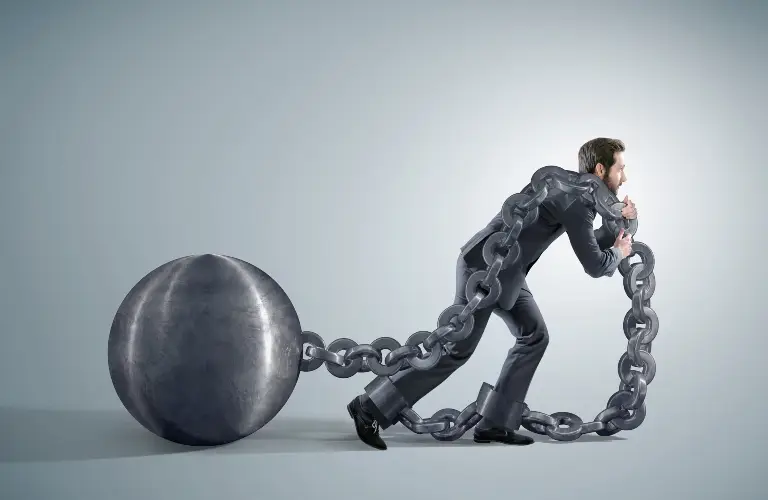 Man in black suit struggling to walk with large metal ball and chain attached.