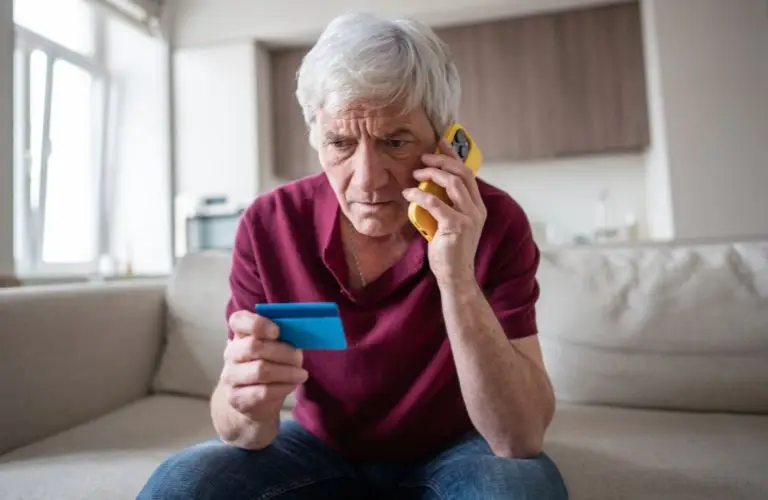 Older man in red shirt sitting on beige couch, holding blue card and yellow phone.